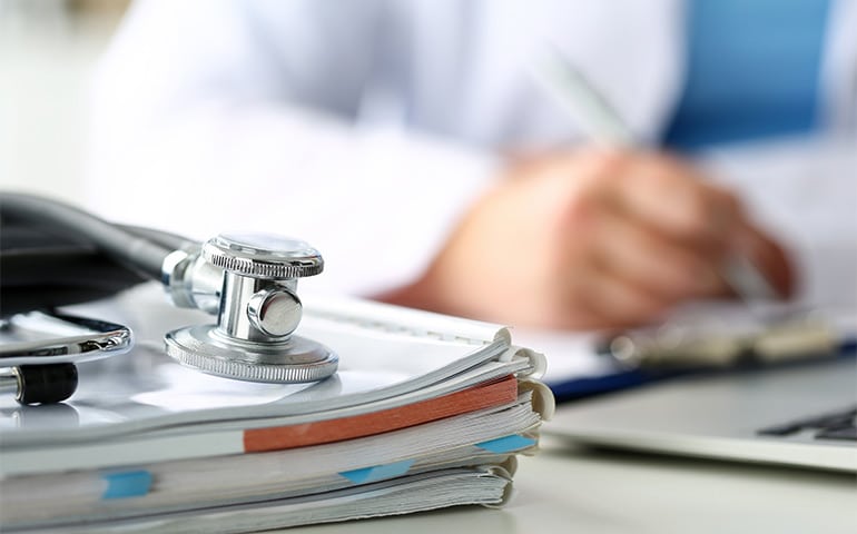 Stethoscope head lying on medical forms on clipboard closeup while medicine doctor working in background. Patient history list, visit check, 911, medical calculation and statistics, healthy lifestyle concept