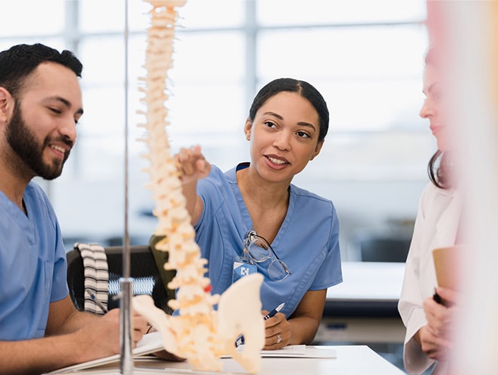 The young adult female medical student asks the mid adult female teacher a question about the human spine as her young adult male friend listens.